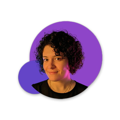 foto de uma mulher branca de cabelo castanho cacheado curto usando uma camiseta preta, colares e piercings dourados, ela olha para frente e está posicionada na frente de dois círculos em tons de roxo